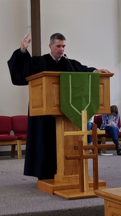Pastor Rob Standing behind a Wooden Pulpit with Three Crosses in Front of it Praying with One Hand Lifted